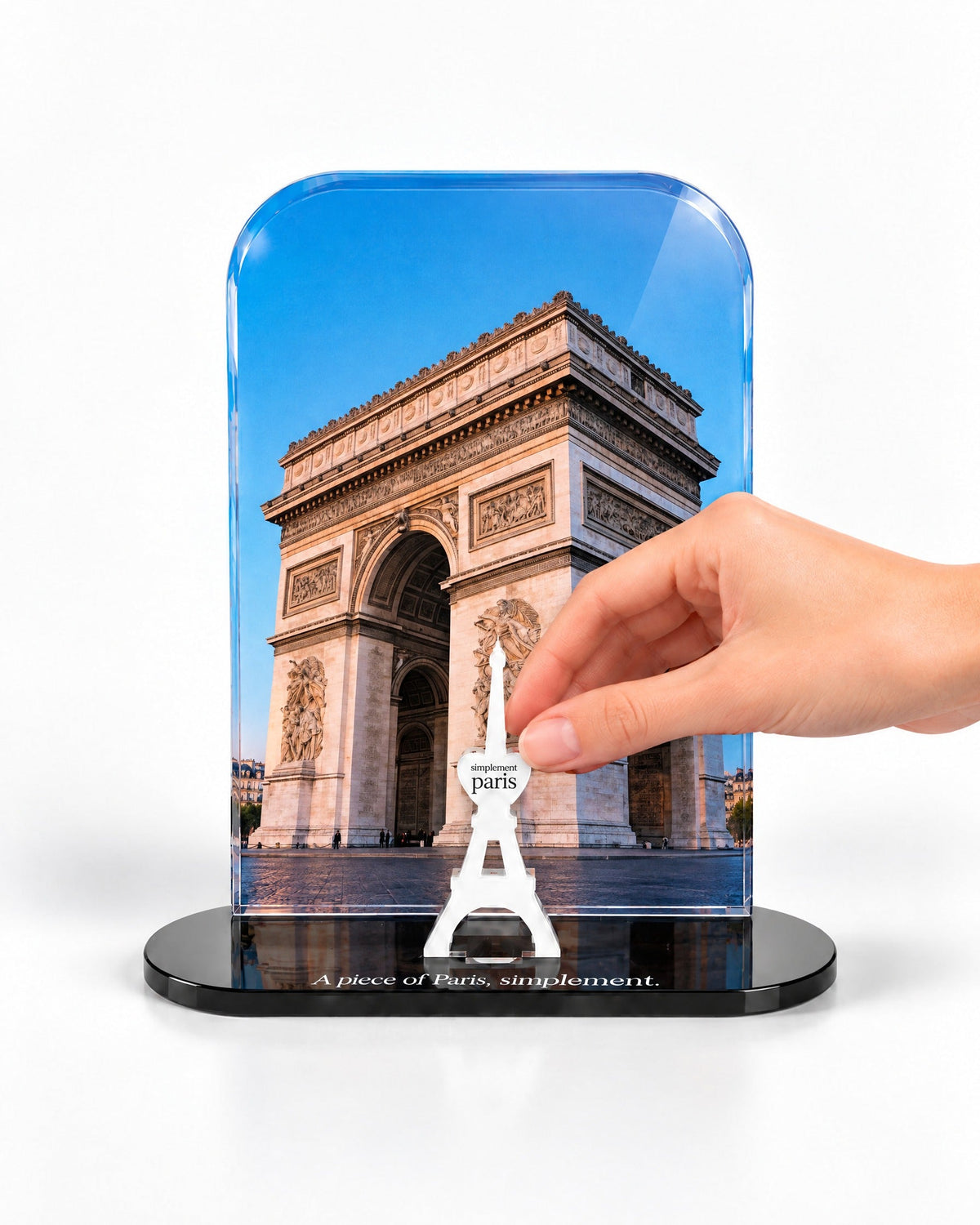 A hand placing the white signature Eiffel Tower in front of the Arc at Dusk acrylic panel