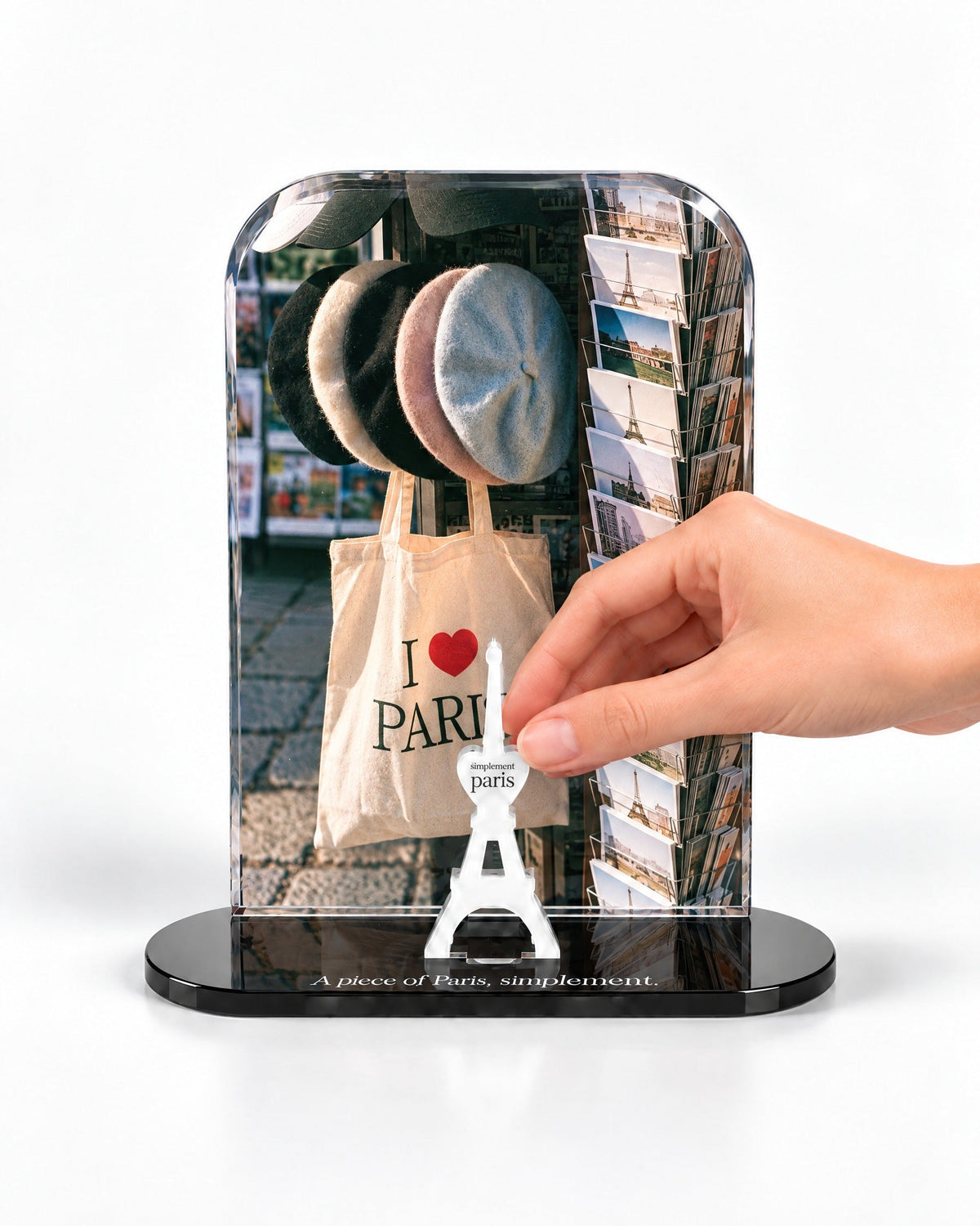 A hand placing the white signature Eiffel Tower in front of the Berets & Postcards acrylic panel