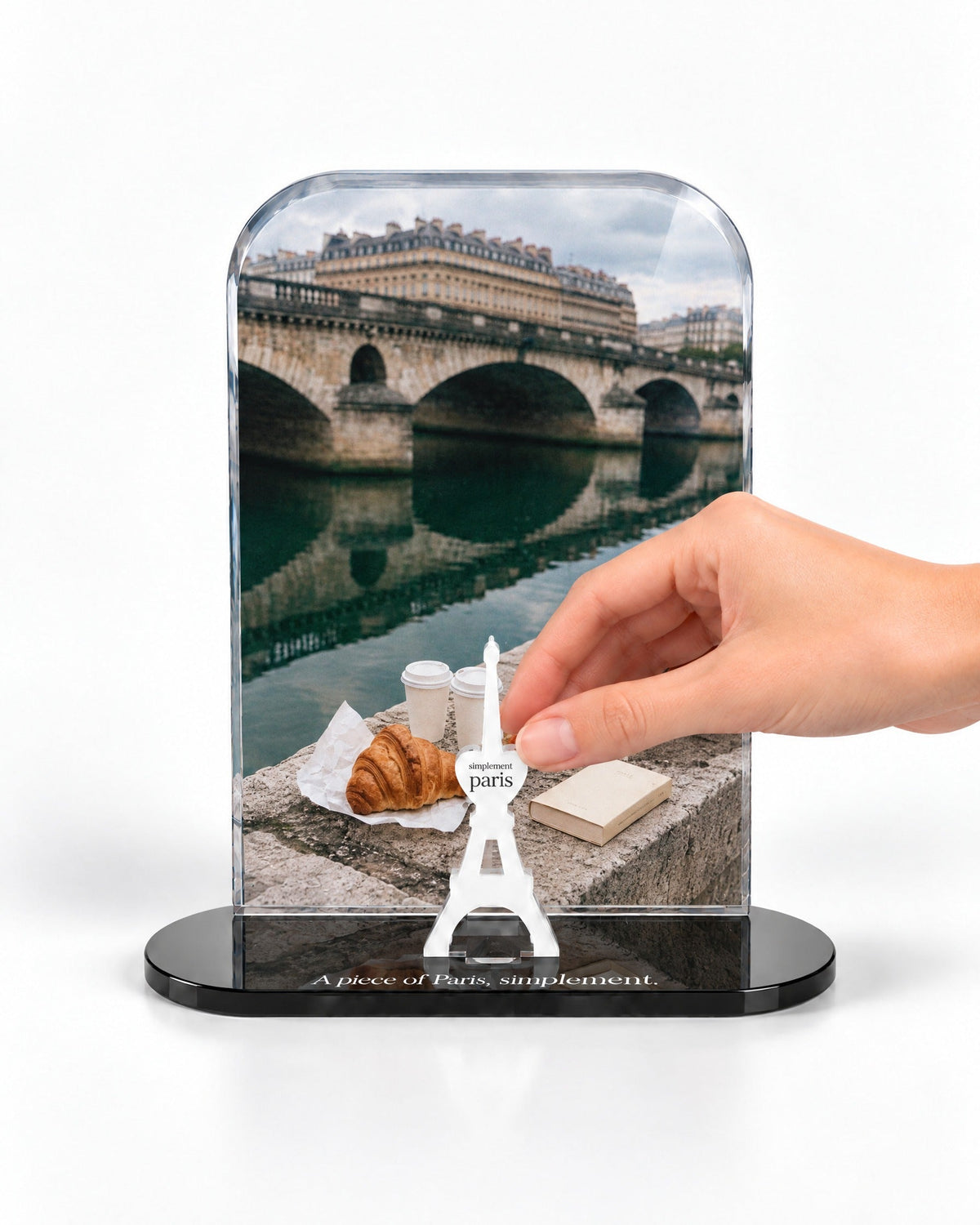 A hand placing the white signature Eiffel Tower in front of the Croissant by the Seine acrylic panel