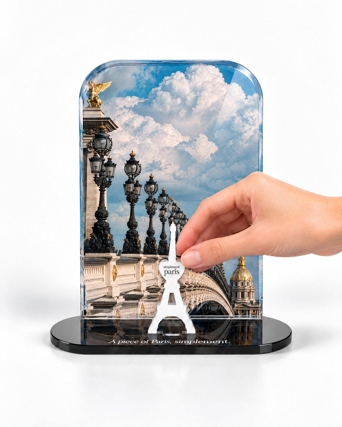 A hand placing the white signature Eiffel Tower in front of the Pont Alexandre III acrylic panel